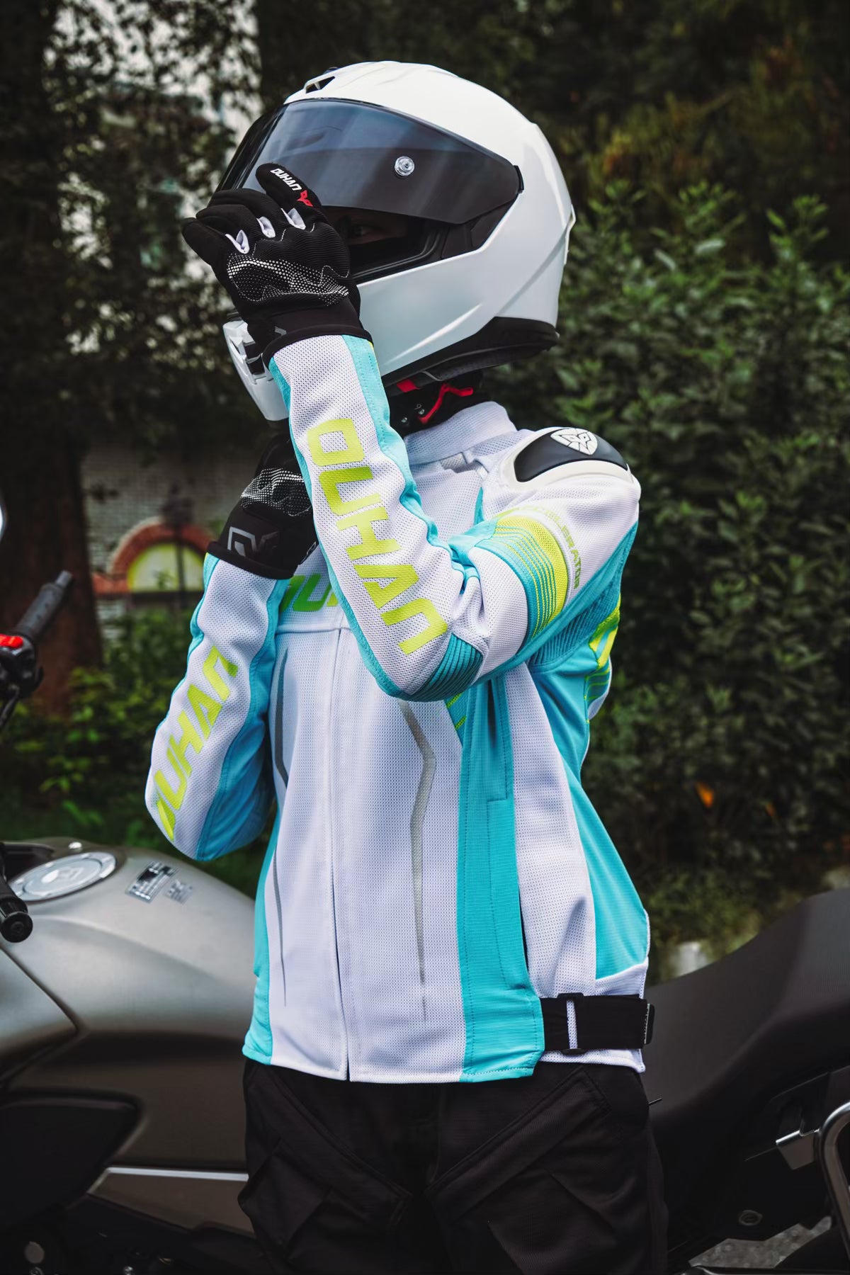 Person wearing a white and blue motorcycle jacket with gloves and helmet, standing next to a motorcycle.